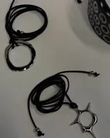 Gargantilla tipo choker de cordón negro, con tres vueltas finas y un dije colgante de metal plateado con forma abstracta o de sol estilizado.