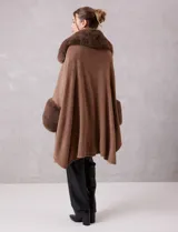 Ruana de tejido color tostado con cuello, bordes frontales y puños amplios con apliques de piel sintética al tono. Presenta una abertura central con cierre de botón tipo trenca.