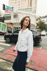 Camisa de popelín blanca con cuello solapa y manga larga. Presenta un diseño frontal con bordados en relieve y botones decorativos negros. Cierre frontal con botones ocultos.