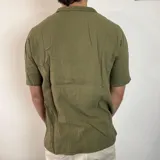 Camisa de hombre color verde oliva, de muselina con textura arrugada, manga corta, corte oversize, cuello cubano y cierre con botones. Presenta un pequeño logo bordado en el pecho.