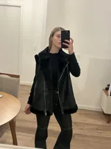 Chaleco negro con cuello solapa y borde de piel sintética, cierre frontal con cremallera y bolsillos laterales con cremallera.