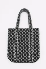 Bolso tipo tote bag color negro con estampado floral blanco, diseño acolchado y asas de hombro con detalles a rayas.