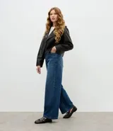 Mocasín femenino de cuero sintético color marrón oscuro, con punta redondeada y suela plana. Presenta un diseño con tiras cruzadas en el empeine decoradas con ojales metálicos y una hebilla ajustable.