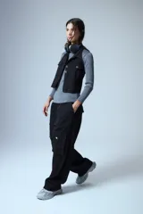 Chaleco de denim negro con escote halter, cuello camisero y cierre frontal con botones metálicos. Presenta dos bolsillos frontales con solapa y diseño sin mangas.