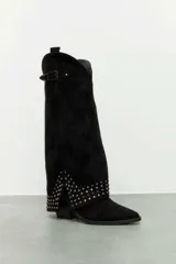 Bota de caña alta estilo slouchy confeccionada en gamuza sintética de color negro, con punta fina y taco bajo. Presenta solapa superior plegada con apliques de tachas metálicas plateadas y una tira con hebilla decorativa en la parte superior de la caña.
