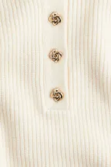 Buzo de punto acanalado color beige, con cuello polo y tapeta frontal con botones metálicos decorativos. Presenta hombros caídos, mangas largas con puños ajustados y diseño de corte recto.