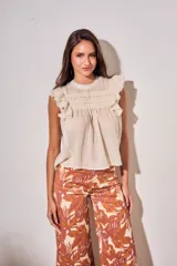 Blusa sin mangas de color crudo, con diseño oversize, cuello redondo y detalles de nido de abeja bordado en el pecho, acompañado de volados en los hombros.