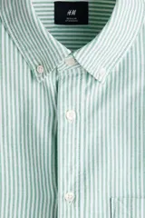Camisa H&M de algodón Oxford con estampado de rayas verticales blancas y verdes, cuello americano, bolsillo frontal de parche y cierre central de botones.