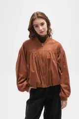 Chaqueta tipo bomber color tostado, confeccionada en material efecto gamuza. Presenta diseño con frunces en el pecho y espalda, cuello mao, cierre frontal completo y mangas largas con puños elásticos.