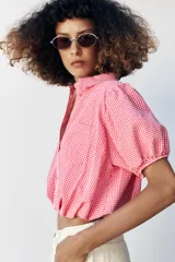 Camisa corta de cuello solapa y manga corta abullonada acabada en elástico, color verde con estampado de cuadros vichy. Cuenta con un bolsillo de plastrón en el delantero, bajo con elástico y cierre frontal con botones.