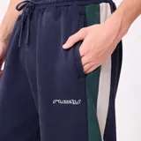 Pantalon de jogging azul marino con franjas laterales en verde y blanco, cintura elástica con cordón ajustable y logo de la marca bordado en la pierna.