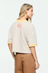 Remera de corte amplio, color beige, con mangas cortas y cuello redondo. Presenta un estampado frontal con la inscripción "The Legendary Hotel Chelsea NY 1884" en tonos celeste y marrón, con detalles en amarillo en el cuello y los puños.