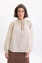 Camisa de manga larga color amarillo pálido con estampa boho a rayas verticales compuestas por pequeños motivos florales. Posee cuello alto con volados y cordones para atar en el frente, y mangas abullonadas con puños ajustados.