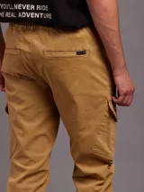 Pantalon tipo cargo color tostado, confeccionado en sarga. Presenta cintura elástica con cordón de ajuste, bolsillos laterales con solapa y puños elásticos en los tobillos.