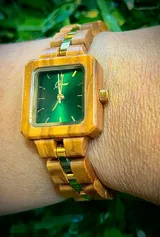 Reloj de pulsera con caja cuadrada de madera de olivo y esfera verde con marcadores minimalistas y agujas doradas. La malla es de madera de olivo con eslabones centrales metálicos en tono dorado.