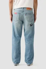 Jean denim de corte holgado, color azul claro, con efecto desgastado y roturas. Presenta parches de tela estampada en rojo con puntos blancos, estampado de vaca en blanco y negro, y un parche amarillo a cuadros.