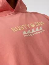 Canguro con capucha color salmón/coral desgastado, con efecto lavado. Presenta un estampado frontal con el texto "RUSTY & SUN" y un diseño de olas en color amarillo pálido. Tiene bolsillo canguro frontal y puños y cintura acanalados.