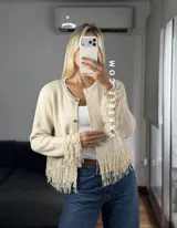 Cárdigan de tejido de punto color crema, con cuello redondo, mangas largas y cierre frontal con botones metálicos. Presenta detalles de flecos en los puños y en el ruedo.