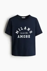 Remera de manga corta color azul marino con cuello redondo y estampado frontal en blanco que dice "MILANO" en arco, "Via della Moda" en letra cursiva pequeña debajo, y "AMORE" centrado abajo.