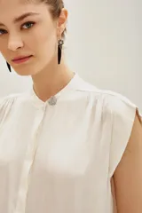 Camisa de corte oversize color negro, con cuello mao, mangas cortas y botones ocultos. Presenta un pin metálico decorativo con strass en el cuello.