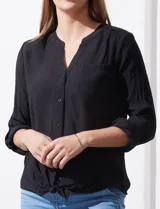Blusa blanca de gasa marca Zac & Rachel, con cuello a la base, escote en V, abertura central con botones, bolsillo aplicado en el pecho y mangas largas regulables con presilla y botón. Tiene terminación opcional de nudo en el ruedo.