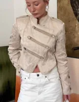 Chaqueta de estilo militar confeccionada en tejido jacquard color beige con sutil brillo. Presenta cuello mao, cierre frontal cruzado con botones decorativos, hombreras con detalles de botones y puños con botones. Posee un calce entallado y largo a la cintura.