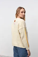 Sweater de punto color crudo con diseño de flores y formas geométricas bordadas en colores vibrantes. Presenta cuello redondo, mangas largas con detalles de líneas laterales en contraste y corte holgado.