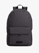Mochila Tommy Hilfiger de diseño minimalista y silueta tipo domo en color gris oscuro. Cuenta con compartimento principal con cierre de cremallera, un bolsillo exterior frontal con solapa y cierre magnético, correas de hombro acolchadas y ajustables, y un asa superior. Presenta el logo de la marca en relieve en el frente y detalles distintivos.
