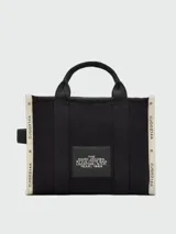 Bolso tipo tote bag de Marc Jacobs en color beige, confeccionado en lona resistente. Presenta el texto "THE TOTE BAG" y "MARC JACOBS" impreso en la parte frontal en color negro. Cuenta con asas superiores y una correa de hombro ajustable y extraíble, además de ribetes laterales negros con inscripciones.