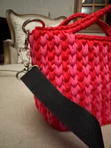 Cartera tejida a crochet con hilo de algodón en tonos rosa y rojo. Tiene doble asa corta y correa larga de tela negra regulable. Incluye un dije de borla rosa.