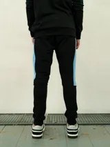 Pantalón de felpa negro con puños elásticos en los tobillos, y una franja lateral azul celeste. Incluye el logo de Umbro en la parte superior del muslo.