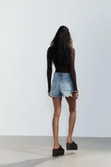 Short de jean negro desgastado, tiro alto, con roturas pronunciadas y terminación deshilachada en el bajo. Presenta cinco bolsillos y cierre frontal con botón metálico.