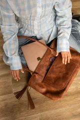 Cartera tipo tote de cuero marrón con costuras artesanales visibles, bolsillo frontal con el logo grabado, asas de hombro y detalles de flecos colgantes.