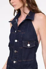Enterito largo confeccionado en denim azul, sin mangas, con cuello halter y solapas de camisa, cierre frontal con botones metálicos y bolsillos frontales con solapa. Presenta pantalón de corte amplio.