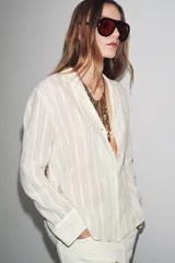 Camisa de algodón color crema o blanco roto, con textura de rayas verticales sutiles. Presenta cuello solapa, escote pico y manga larga.