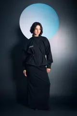 Campera negra de cuello alto con cierre frontal de botones a presión, mangas largas y cordones ajustables en la cintura. Presenta costuras a la vista en color blanco y un bordado decorativo en el pecho.