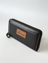 Billetera grande de cuero negro con cierre metálico perimetral, costuras blancas visibles y un parche marrón claro con el logo grabado en el frente.