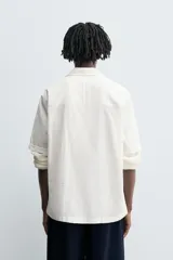 Camisa de calce relajado confeccionada en tejido ligero de algodón con textura. Presenta cuello bowling, manga corta, un bolsillo de parche en el pecho y cierre frontal con botones. El bajo cuenta con aberturas laterales.