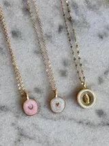 Collar dorado con cadena fina y dije circular con borde dorado y centro de esmalte blanco con figura de la Virgen María en dorado y strass. También disponibles con dije circular con esmalte rosa y letra 'C' dorada, y con esmalte blanco y corazón dorado.