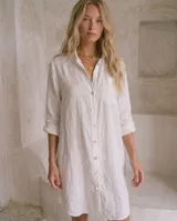 Vestido camisero de lino color blanco, con cuello camisero, cierre frontal de botones, bolsillos en el pecho y mangas tres cuartos con presillas para arremangar. Diseño de corte recto y largo midi.