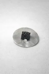 Broche para el pelo con base circular de metal plateado (alpaca o bronce) y una pieza central cuadrada de piedra negra con textura rugosa y cristalina.