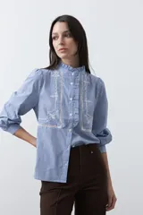 Camisa de rayas verticales con cuello mao, detalle de volados en el escote, bordados florales y recortes con calados. Posee mangas largas abullonadas con puños ajustados y cierre frontal con botones.