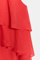 Top de cuello redondo y manga sisa, confeccionado en tejido con acabado satinado. Presenta volantes en cascada y un diseño de bajo asimétrico, con cierre en la espalda mediante pequeña abertura y botón.