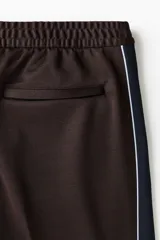 Pantalón deportivo marrón de punto con leve brillo, corte relajado y piernas rectas con pliegues cosidos. Cuenta con franjas laterales oscuras y ribetes blancos contrastantes, cintura elástica y bolsillos laterales.