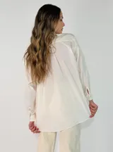 Camisa de corte oversize y diseño holgado, con cuello camisero, cierre frontal de botones y mangas largas con puños abotonados. Presenta un ruedo redondeado y una caída fluida.