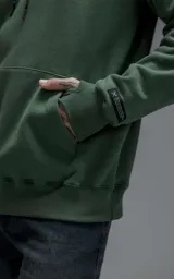 Hoodie oversize color verde, confeccionado en frisa premium con textura gamuzada. Presenta capucha ajustable con cordones personalizados, bolsillo frontal tipo canguro y puños con abertura para pulgar.