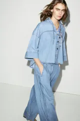 Camisa de denim en tono azul claro, con cuello solapa, escote en pico y cierre frontal con botones. Presenta mangas anchas por debajo del codo y un diseño de bajo asimétrico.