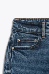 Jeans de mujer, tiro alto, corte Mom Fit con pierna slim y largo tobillero. Denim azul medio con efecto desgastado y lavado. Presenta cinco bolsillos y cierre frontal con cremallera y botón.