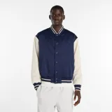 Campera tipo varsity New Balance, con cuerpo en azul marino y mangas en color crema. Presenta cierre frontal con botones a presión, cuello, puños y ruedo acanalados con rayas en contraste, y logo bordado en el pecho.