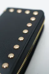 Billetera negra con cierre perimetral y tachas doradas decorativas en el borde frontal.